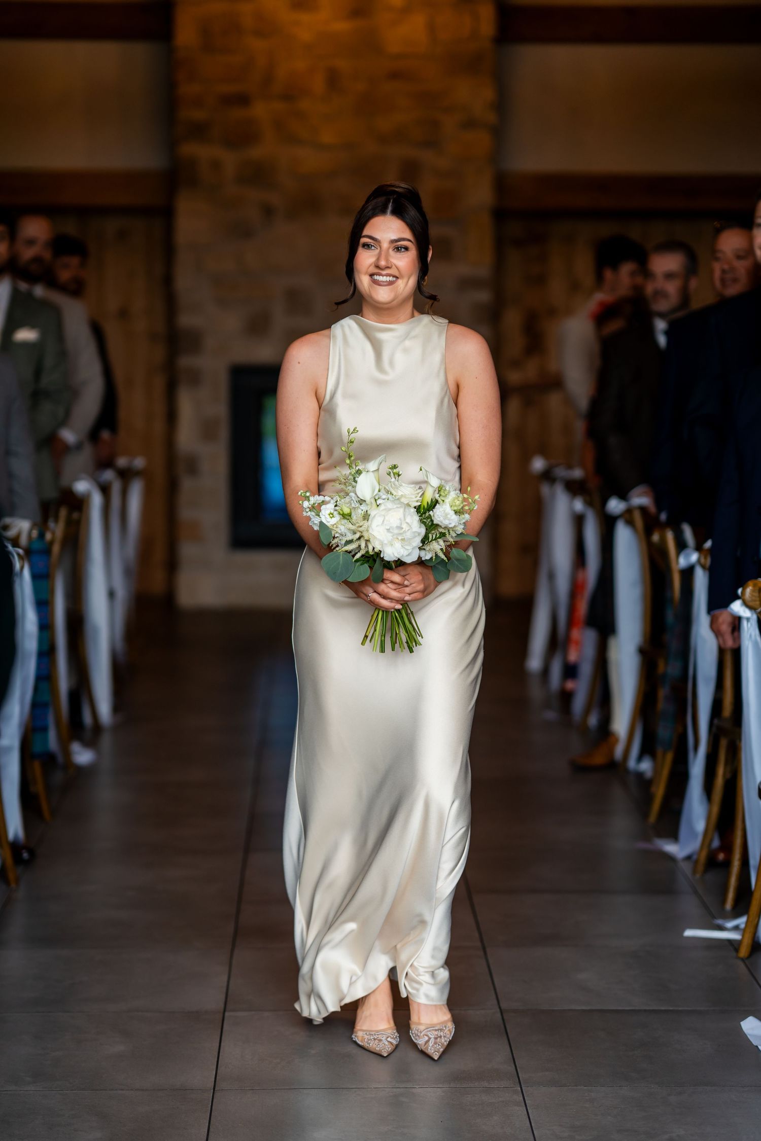 Smiling bridesmaid walks down church aisle in elegant champagne satin gown holding white floral bouquet.