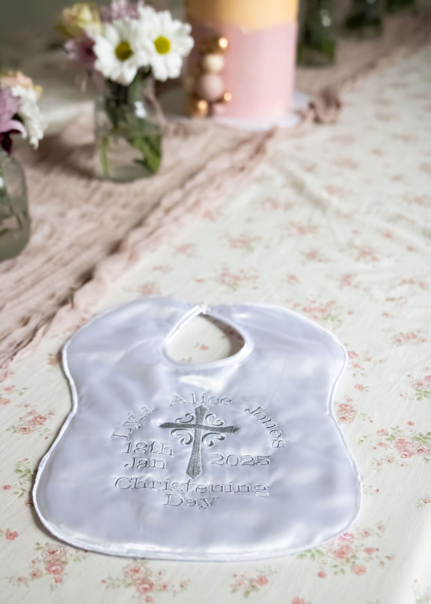 White embroidered baby bib with cross design and christening day date, displayed on floral tablecloth.