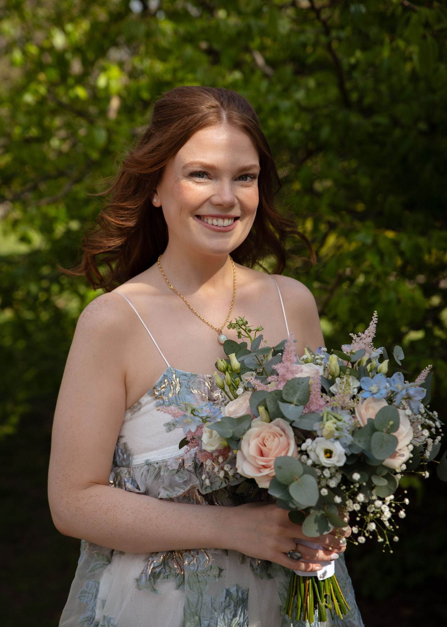 A bride in a light blue floral dress holding a pastel bouquet outdoors.