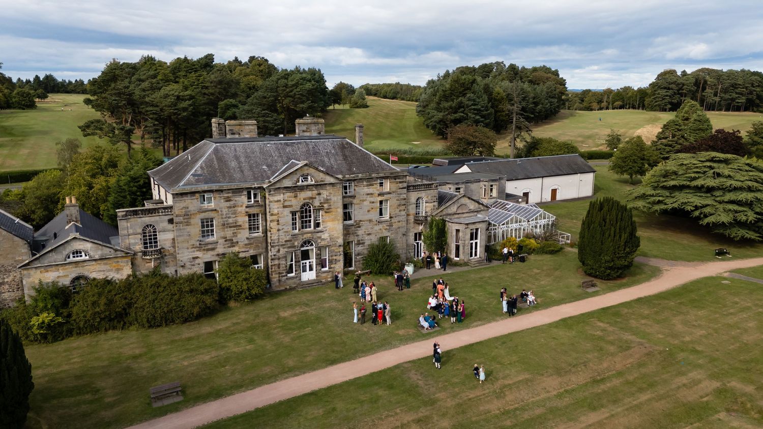 Aerial view of a grand stone country house estate with manicured lawns and guests gathering outside on a sunny day.