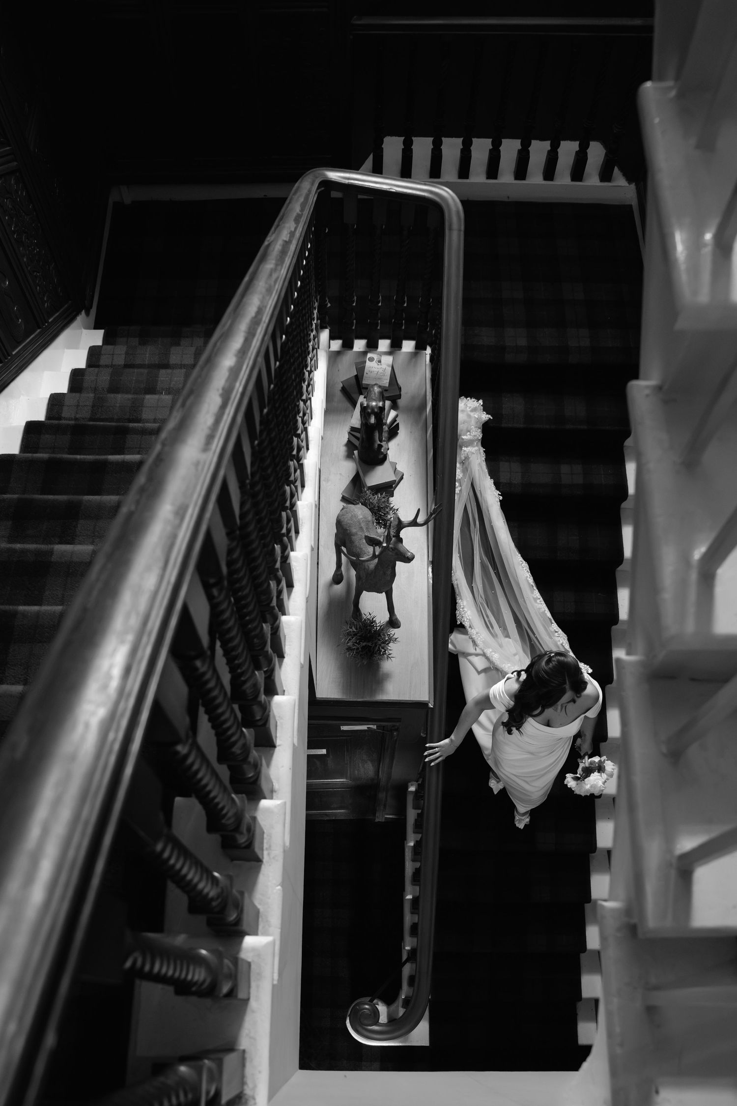 Black and white aerial view of a bride descending an elegant staircase, with wedding guests visible below.