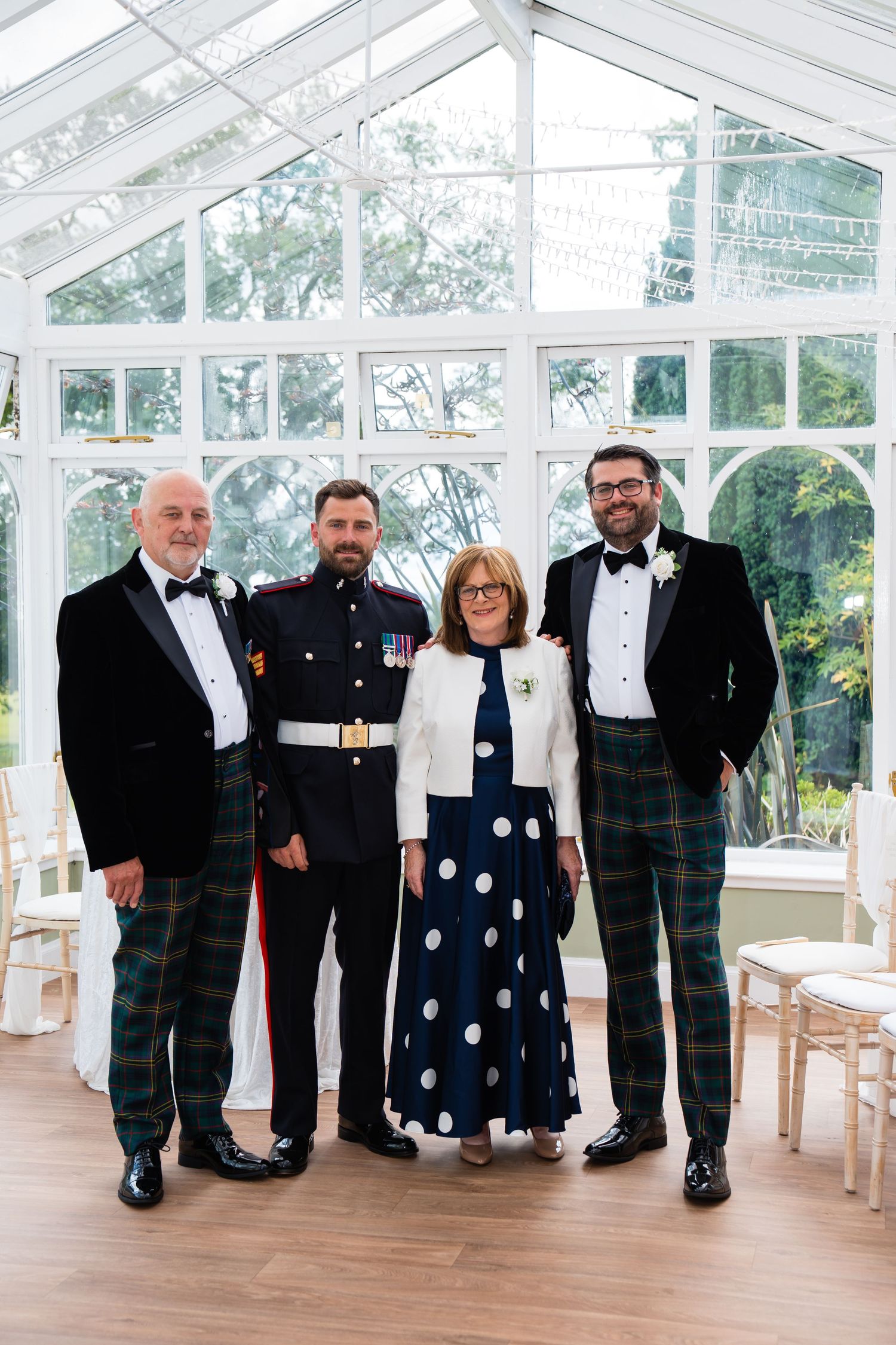 Group of four posing in a glass conservatory, three men in tartan trousers and one woman in a polka dot dress.