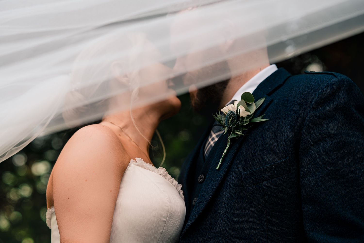 A bride and groom share a romantic kiss under a flowing white wedding veil, groom wearing a navy suit with floral boutonniere.