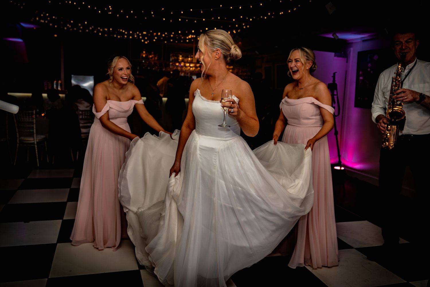 Bride in white gown dancing with bridesmaids in pink dresses on checkered dance floor at wedding reception.