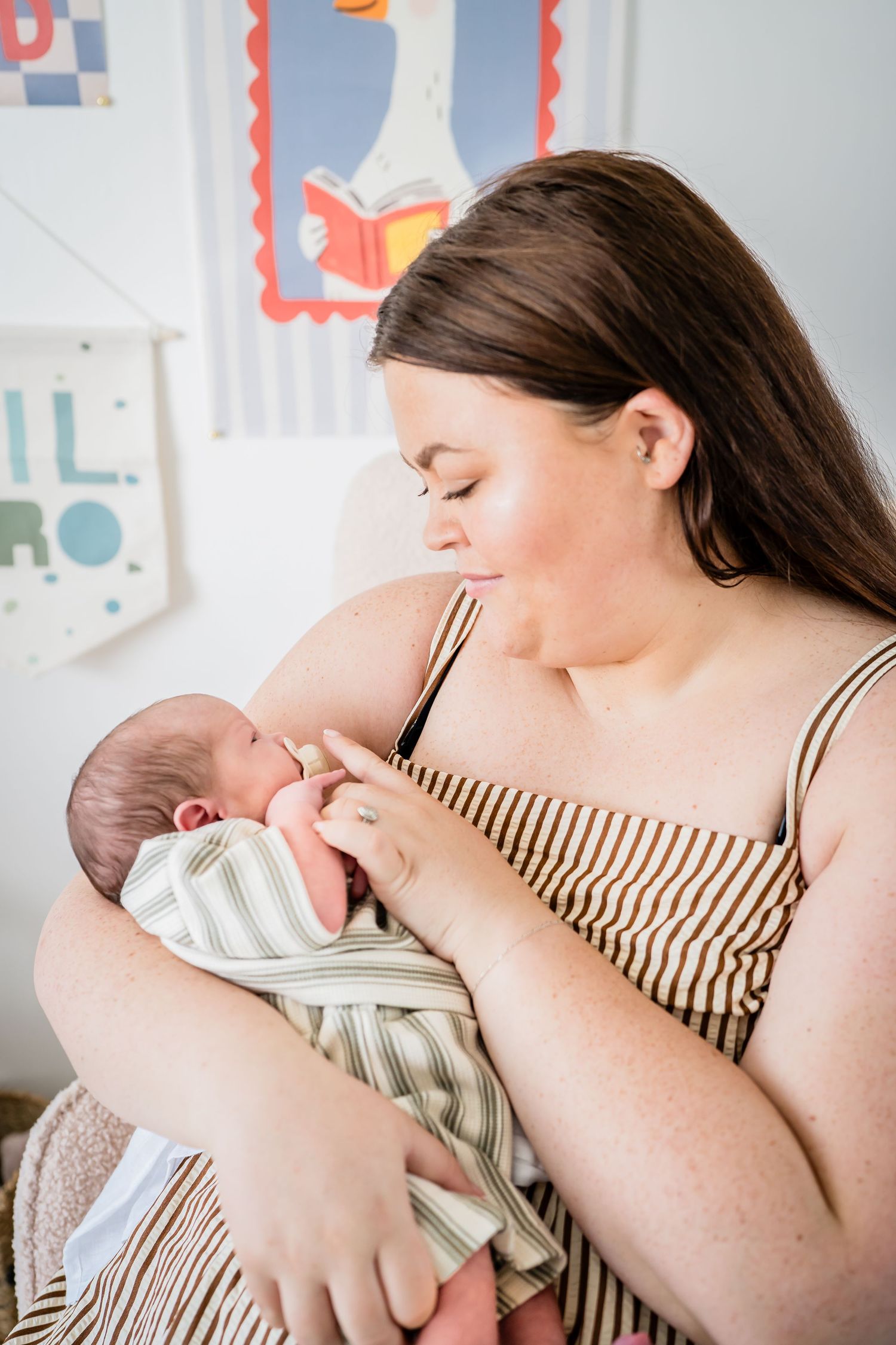 A young mother with long brown hair tenderly cradles her newborn baby wrapped in a green striped swaddle blanket.