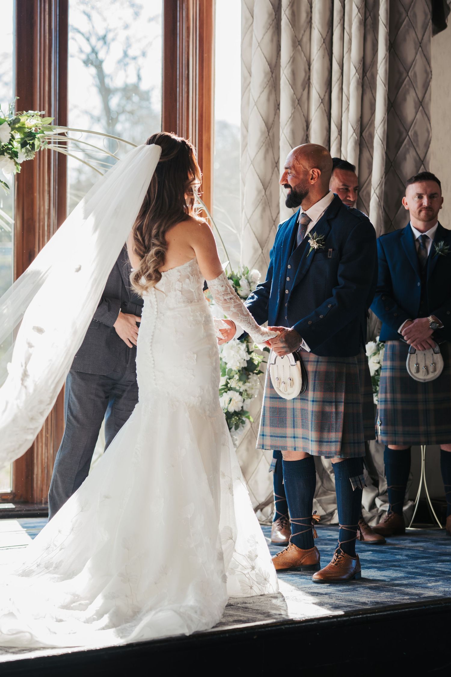 A wedding ceremony shows a bride in a white mermaid gown exchanging vows with groomsmen wearing traditional Scottish kilts.
