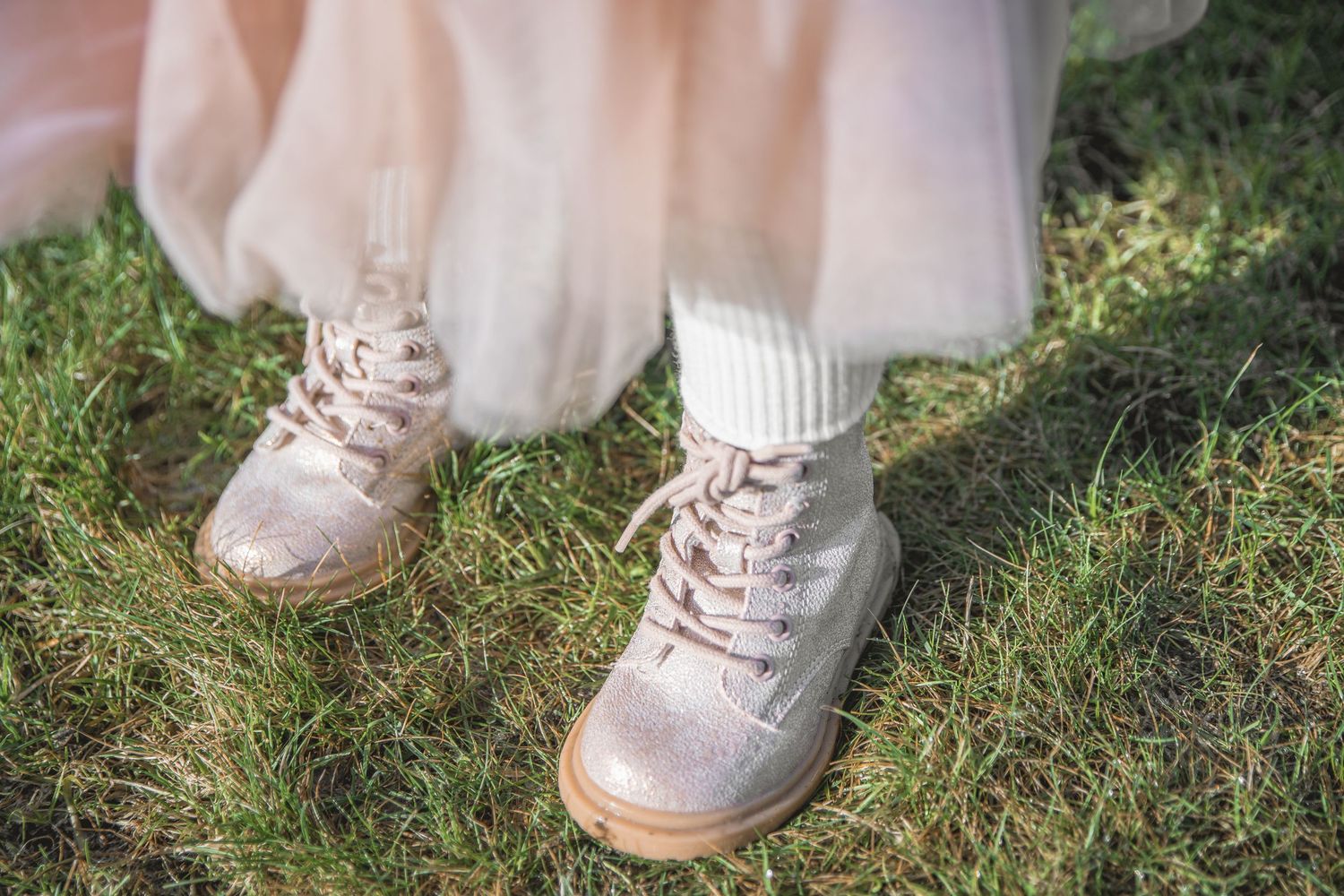 Close-up of pink glitter lace-up boots with white socks worn over green grass with a blush tulle skirt.
