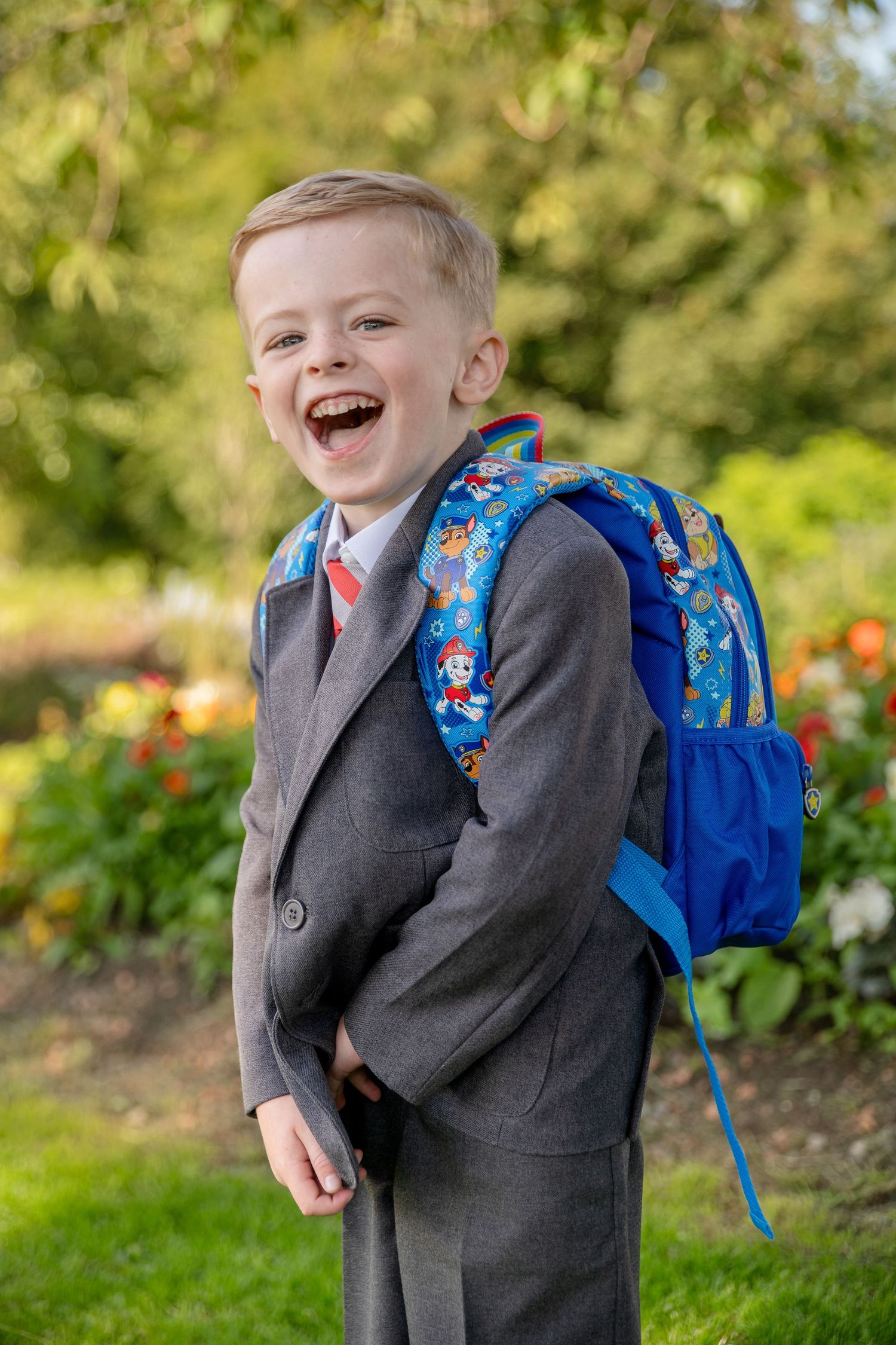A cheerful young boy in a gray suit with a blue backpack laughing outdoors on his first day of school.