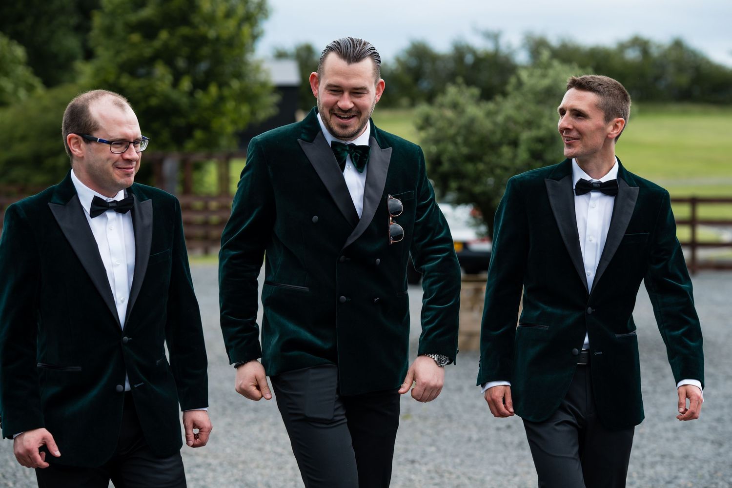 Three groomsmen in green velvet tuxedo jackets and black bow ties walking together outdoors at a wedding.