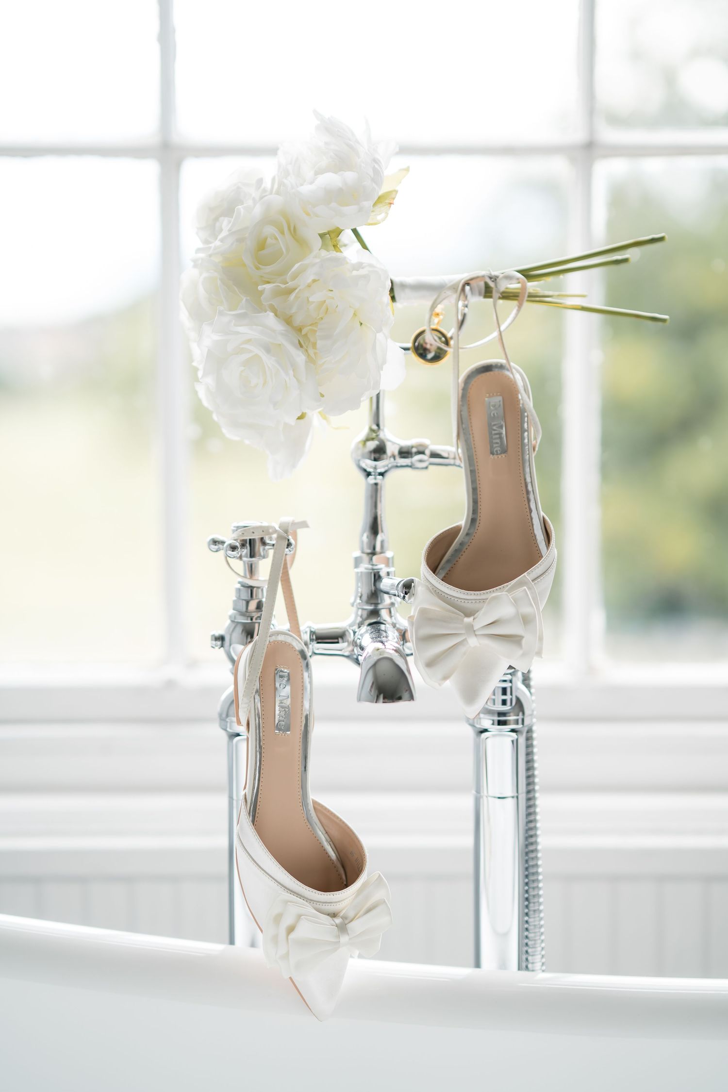 White bow heeled bridal sandals displayed on a vintage chrome bathtub faucet with white roses and gold earrings.