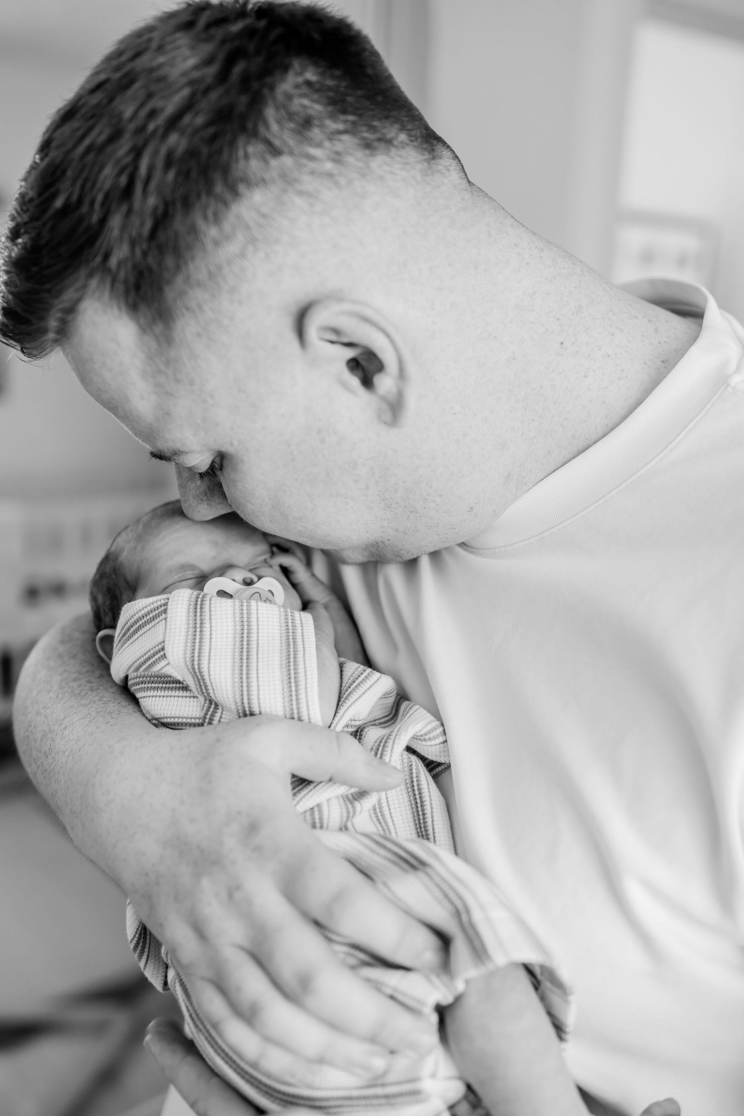 Black and white photo of a father tenderly kissing his newborn baby while cradling the infant in his arms.