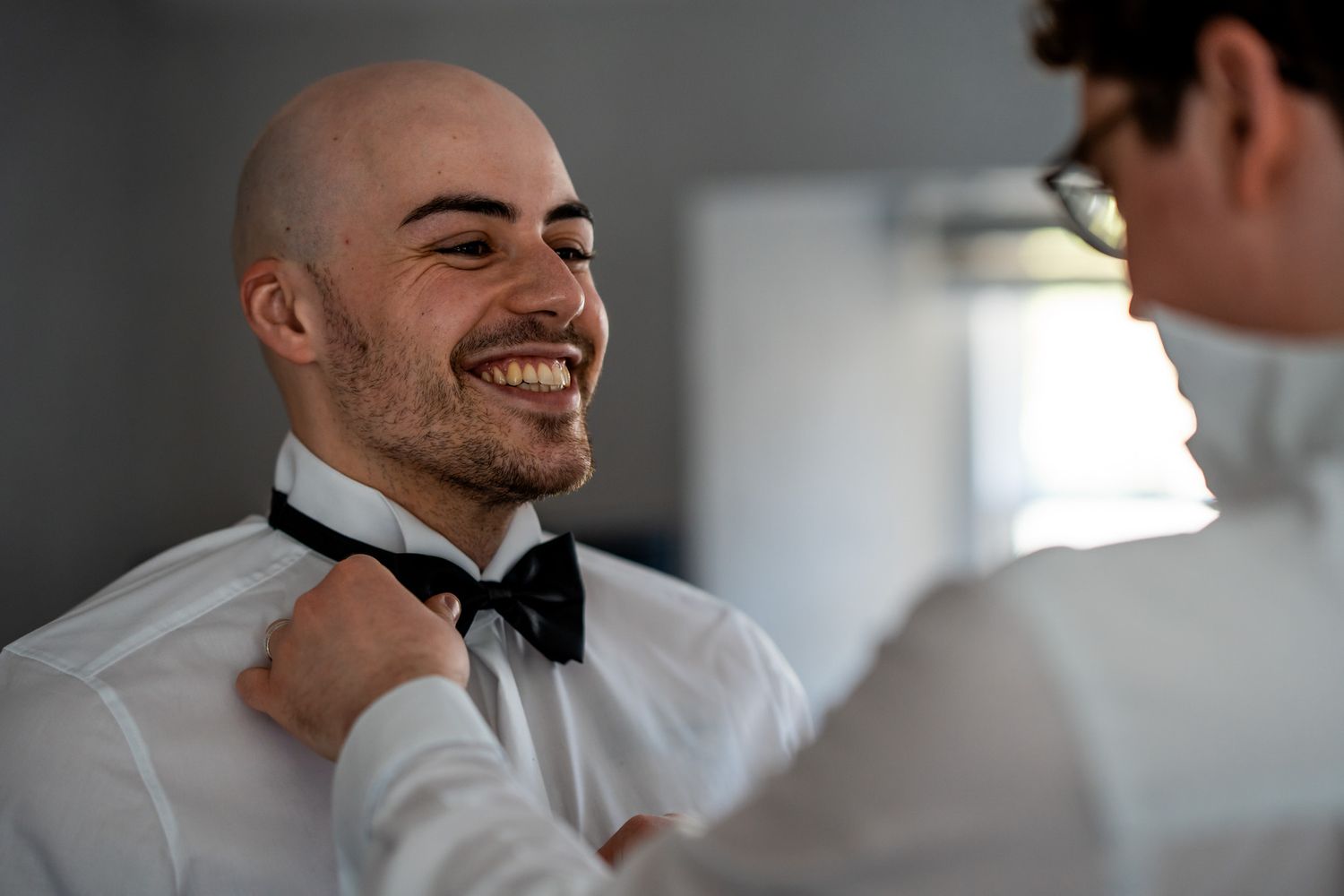 A smiling bald groom having his black bow tie adjusted by a friend while getting ready for his wedding day.