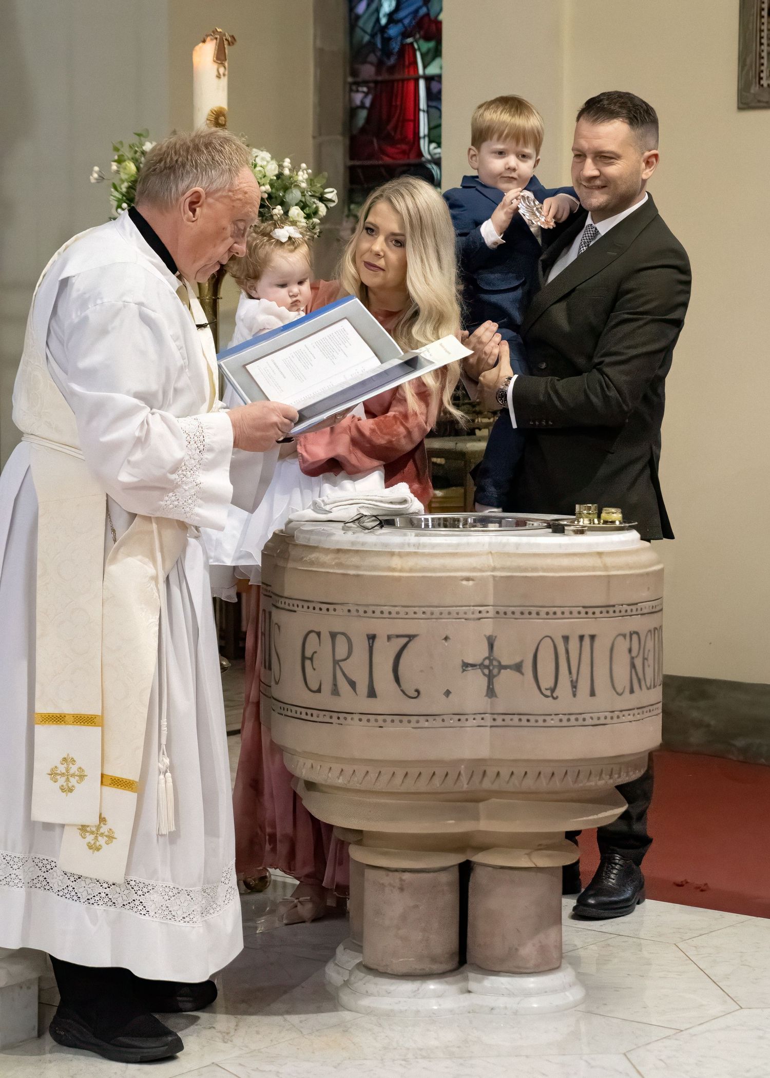 Priest performing a baptism ceremony at a stone font with a family including two young children in a church.