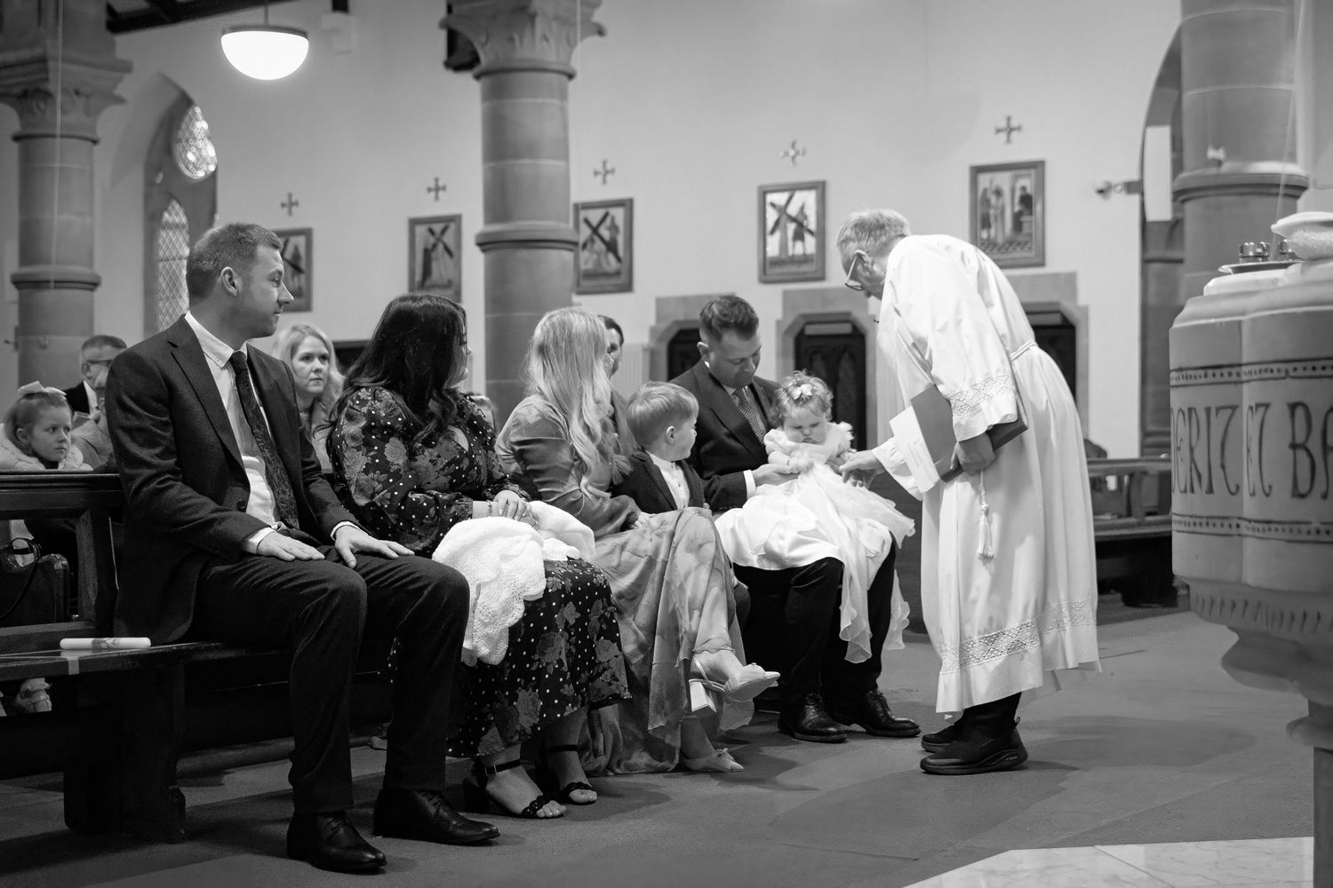 Black and white photo of a priest performing a baptism ceremony with family seated in church pews.