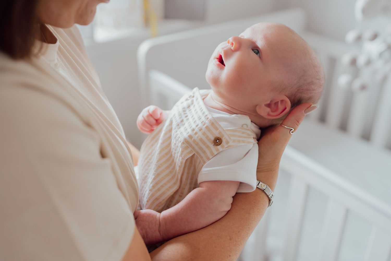 Mother holding a newborn baby in a nursery room with a white crib in the background.