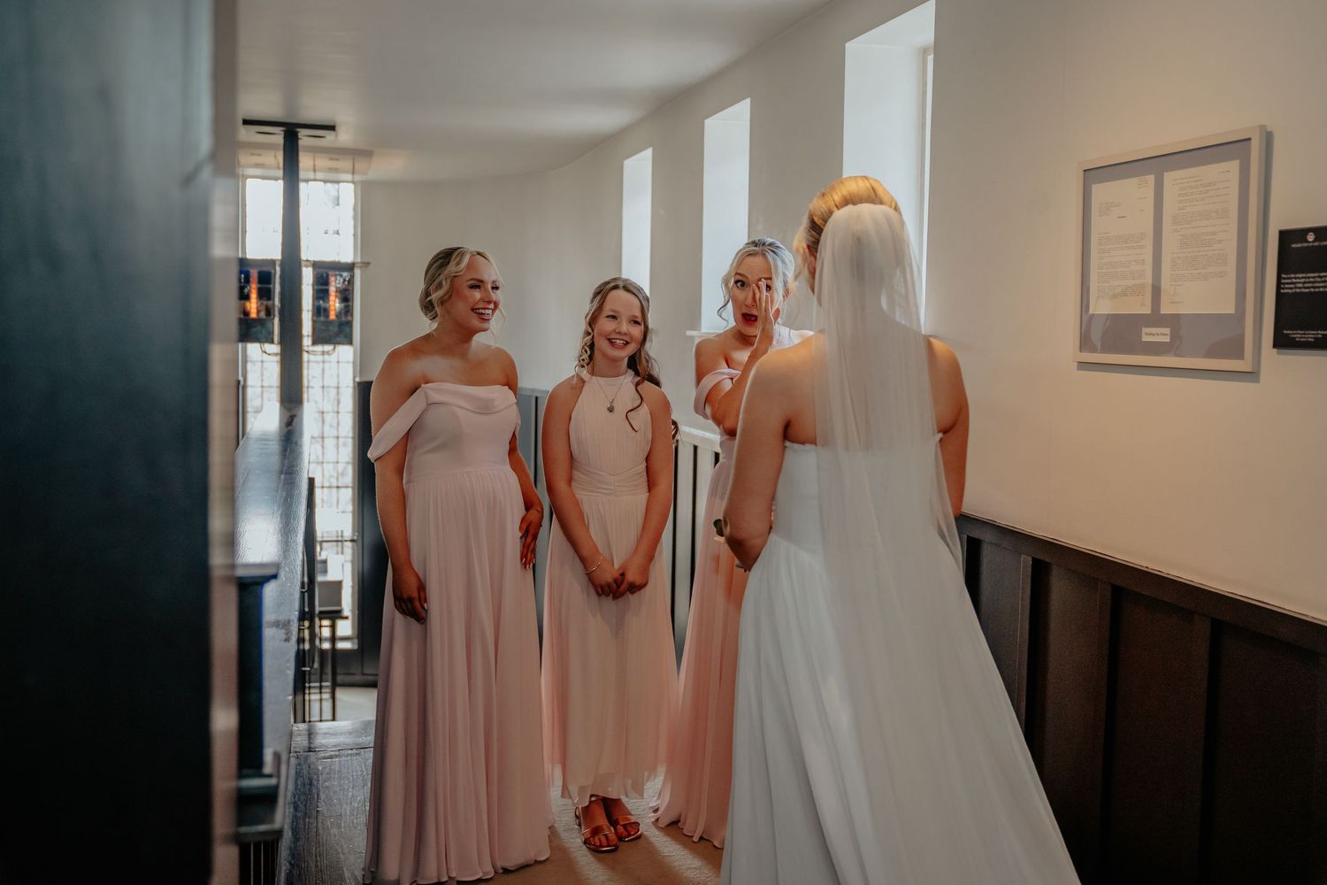 Bride in white gown and veil shares a joyful moment with bridesmaids in blush pink dresses inside a church.