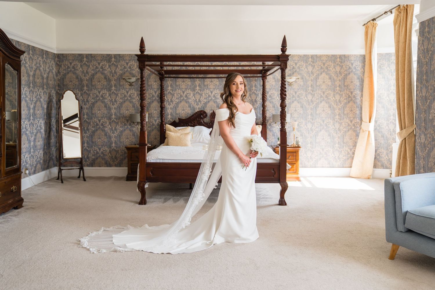 Bride in elegant white gown with long train stands in luxurious four-poster bedroom with damask wallpaper.