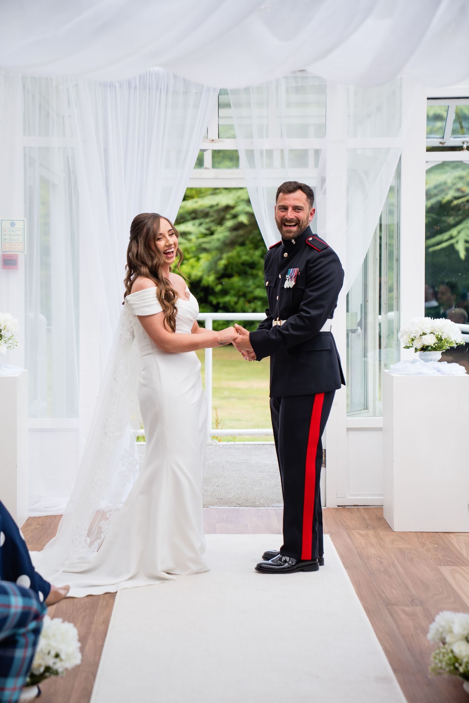 Bride in white gown and groom in military uniform holding hands and smiling at wedding ceremony altar.