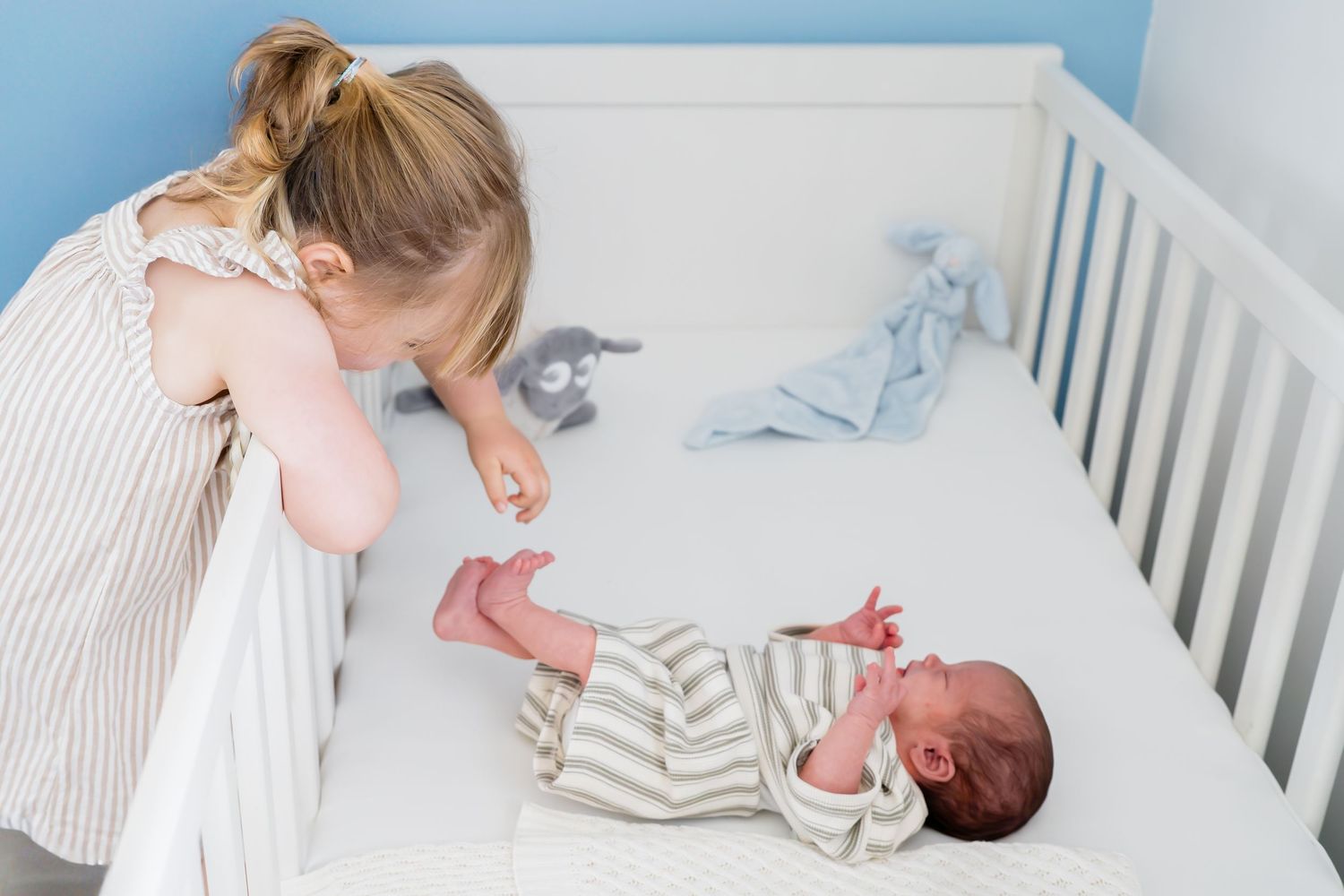 Young girl leaning over white crib looking at newborn baby in striped onesie with blue stuffed animal nearby.