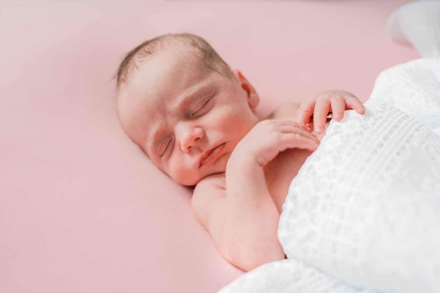 Peaceful newborn baby sleeping on a soft pink blanket, wrapped in a white swaddle with tiny hands visible.