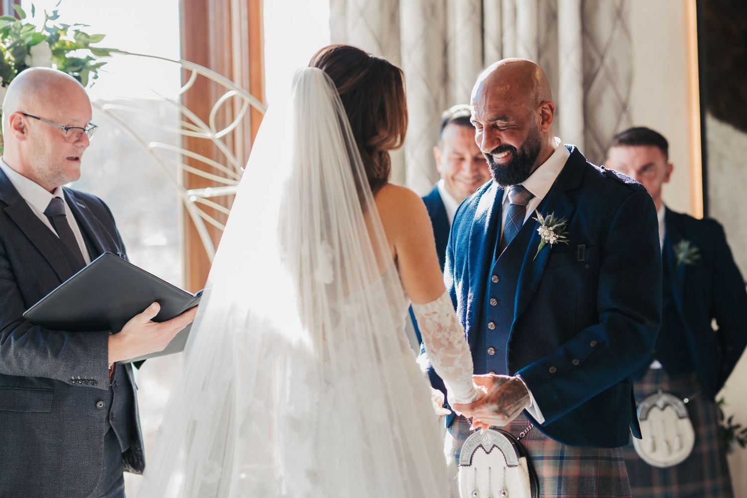 A couple exchanges vows during an indoor wedding ceremony with an officiant and guests looking on.