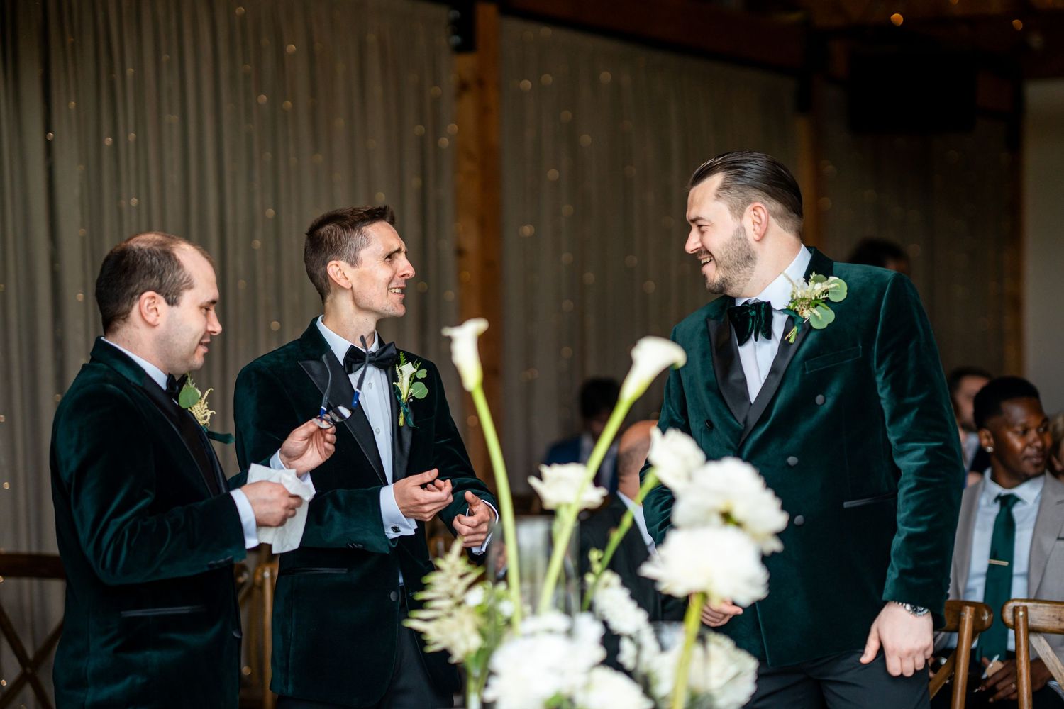 Three groomsmen in green velvet tuxedos with black bow ties chatting at an elegant wedding reception.