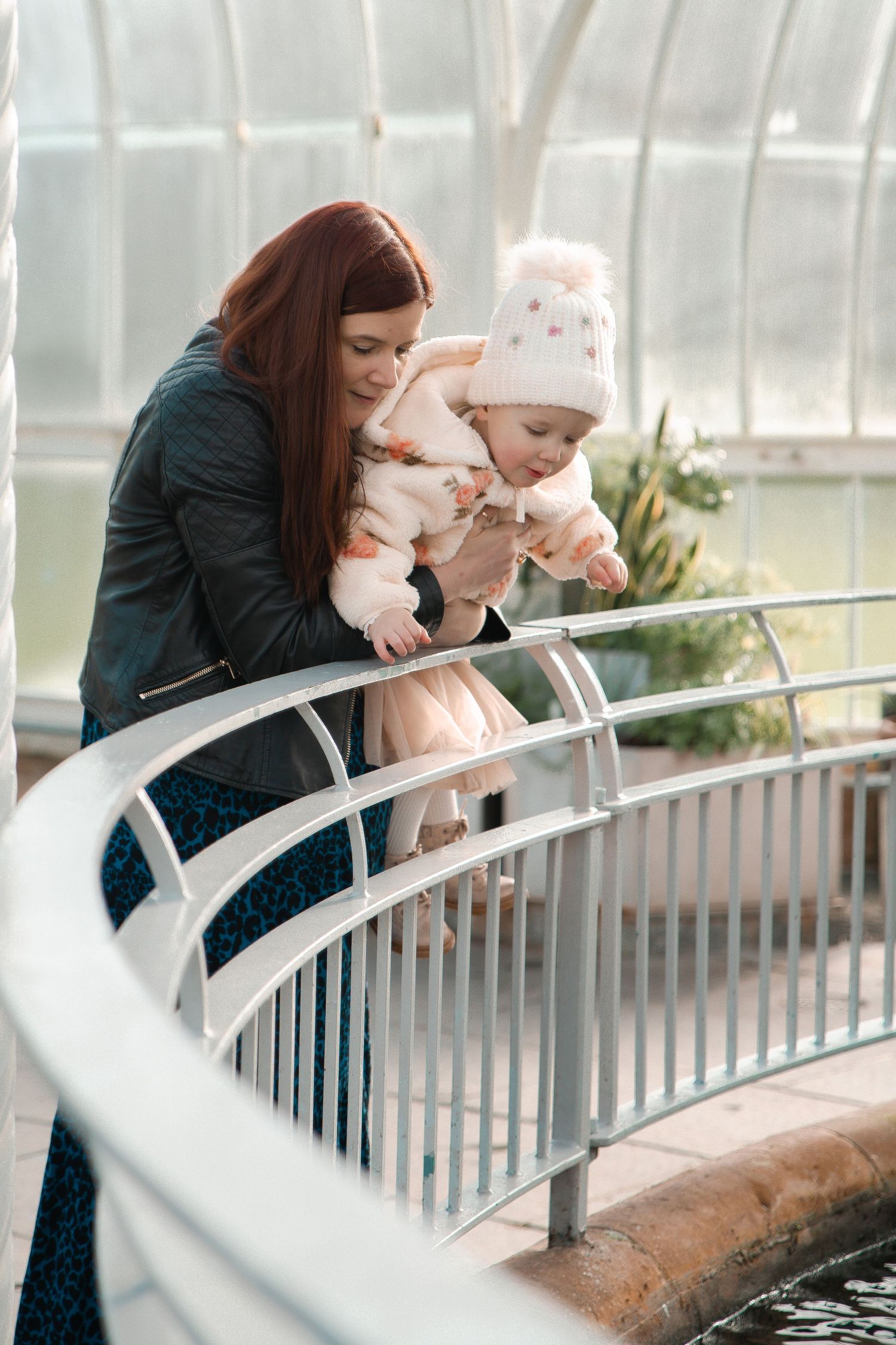 A mother with red hair holds her toddler girl in a white hat on a curved bridge inside a greenhouse.