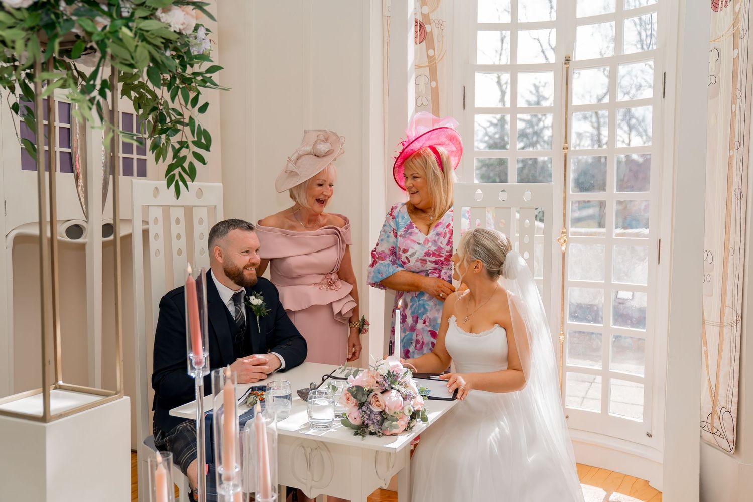 Bride and groom signing wedding register at elegant venue, watched by two guests in pink formal attire.
