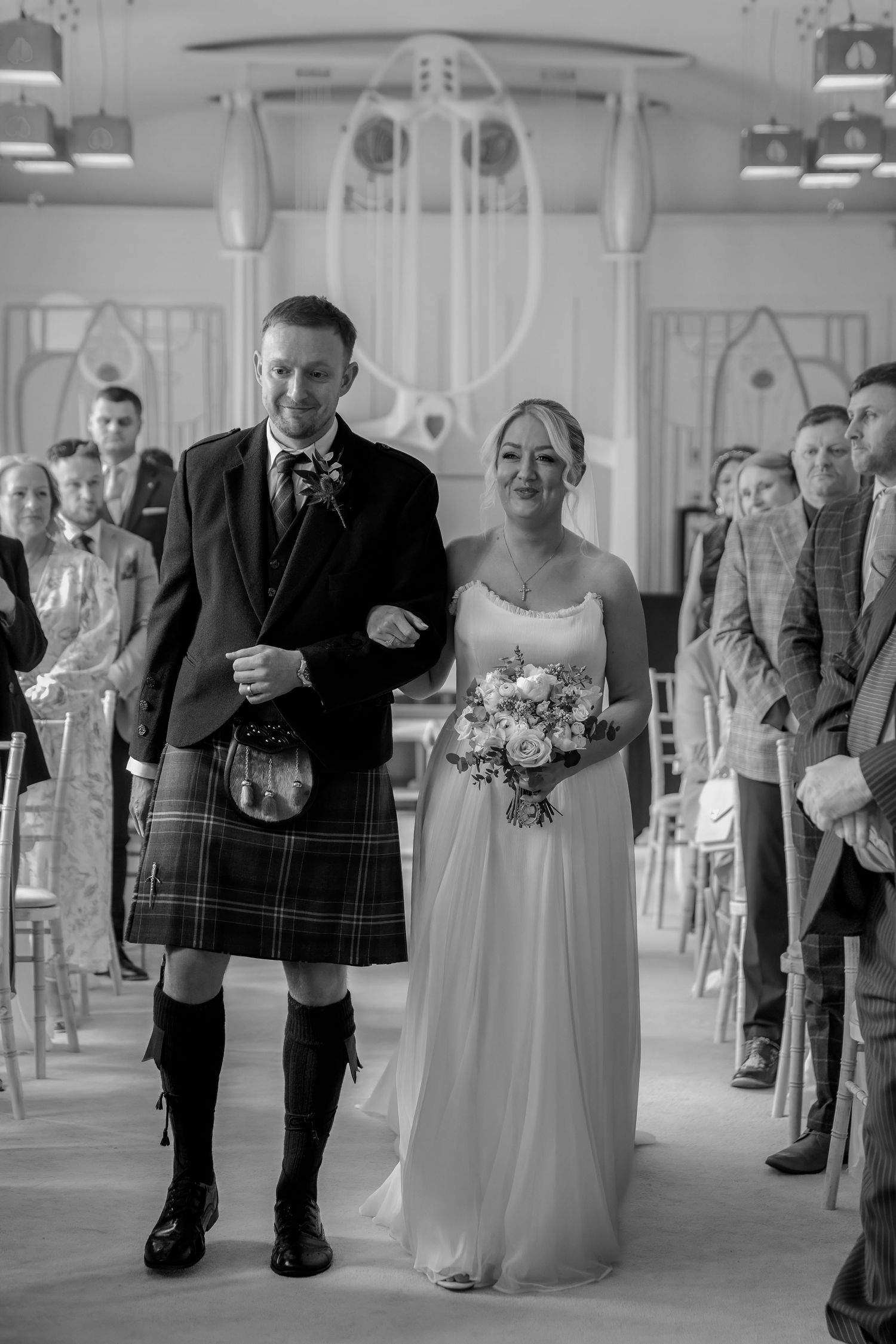 Black and white photo of a groom in a kilt and bride holding bouquet walking down the aisle at a Scottish wedding ceremony.
