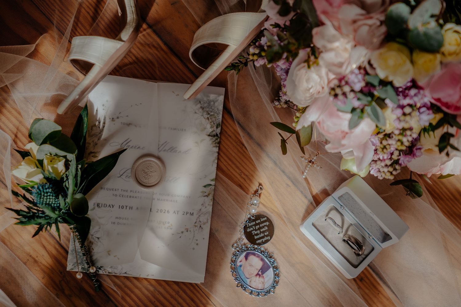 Wedding flat lay with invitation, wax seal, boutonniere, bridal bouquet, earrings, and heels on wooden table.