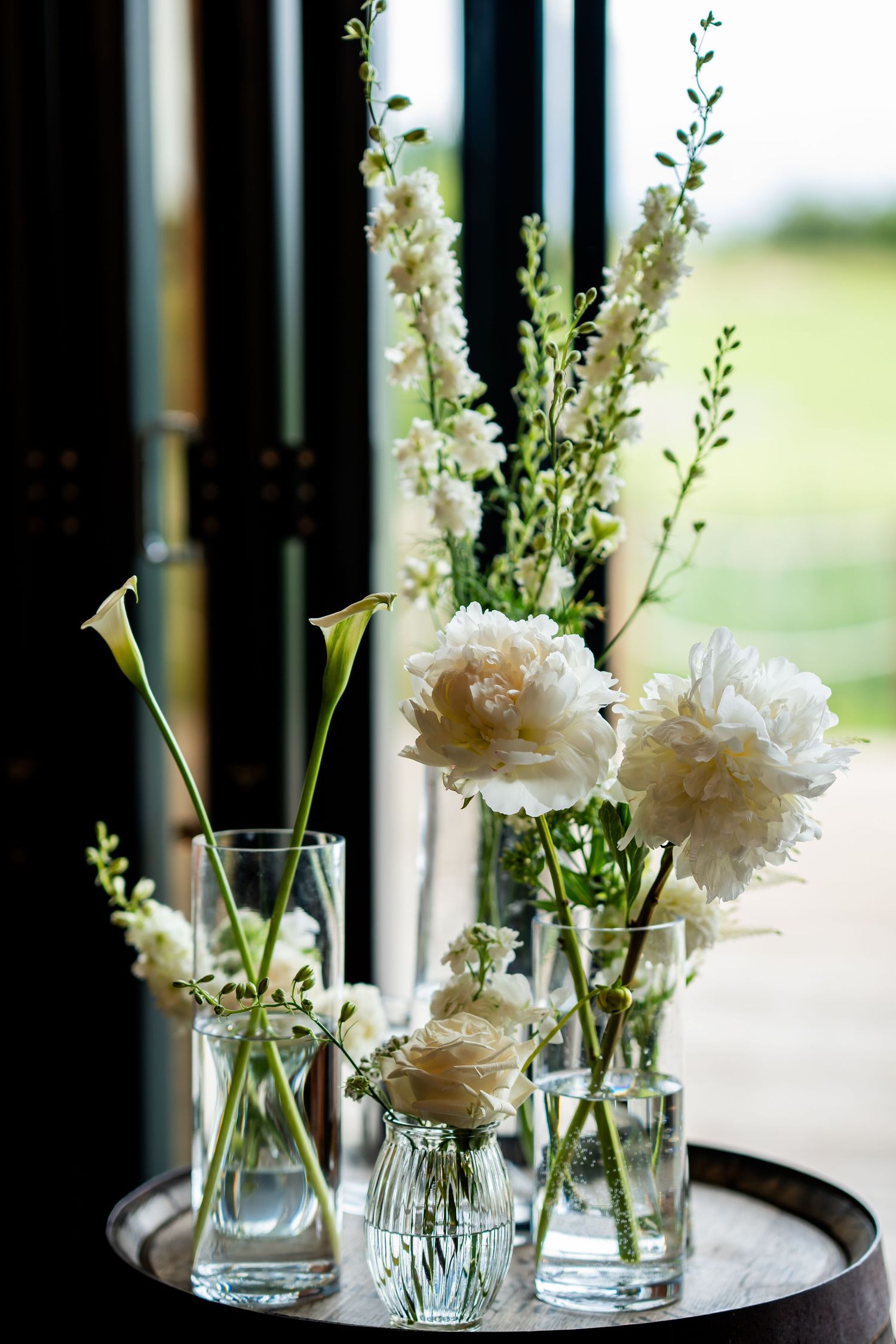 White roses, peonies, and calla lilies arranged in clear glass vases on a silver tray near a window.