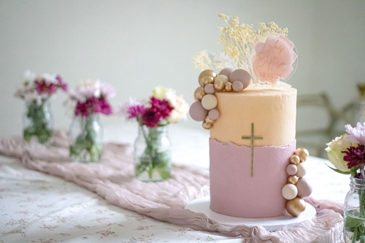 Elegant two-tier pink and gold baptism cake with cross, pearl decorations, and floral topper on a decorated table.