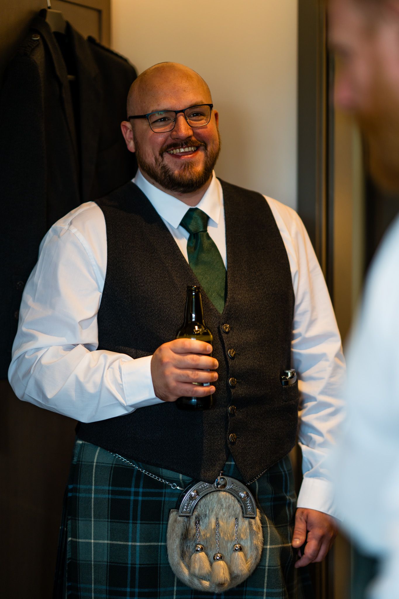 Smiling bald man in Scottish tartan kilt, green tie, and tweed vest holding a beer bottle at a wedding.