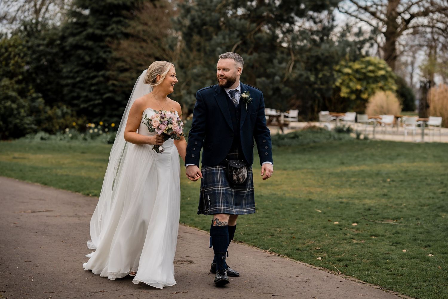 Bride in white gown and groom in blue tartan kilt walk hand-in-hand through a lush green park.