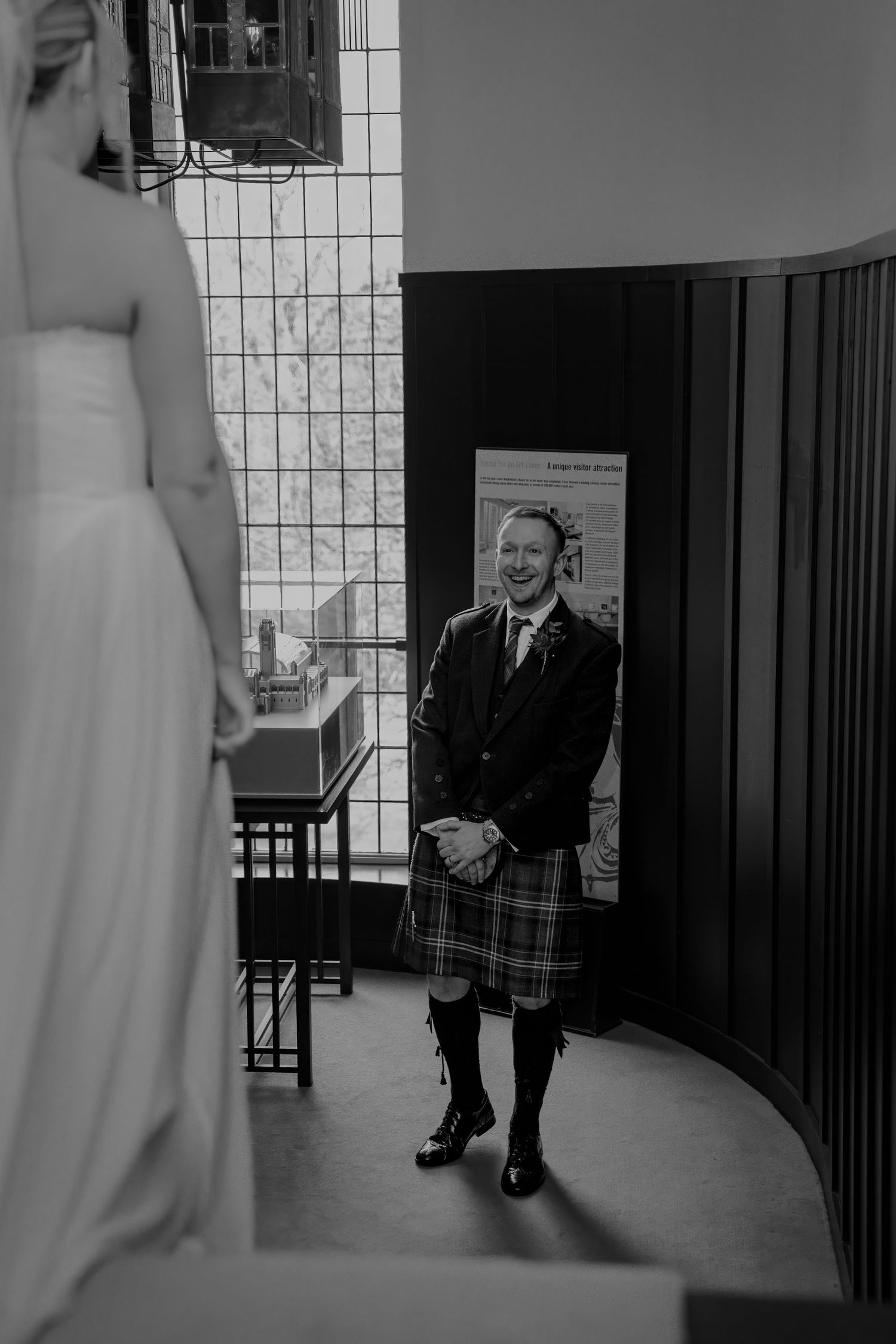 Groom in traditional Scottish kilt smiling at bride during wedding ceremony in black and white photo.