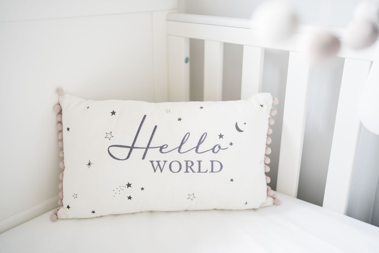 White nursery pillow with Hello World text, stars, and pink pom-pom trim in a white crib.