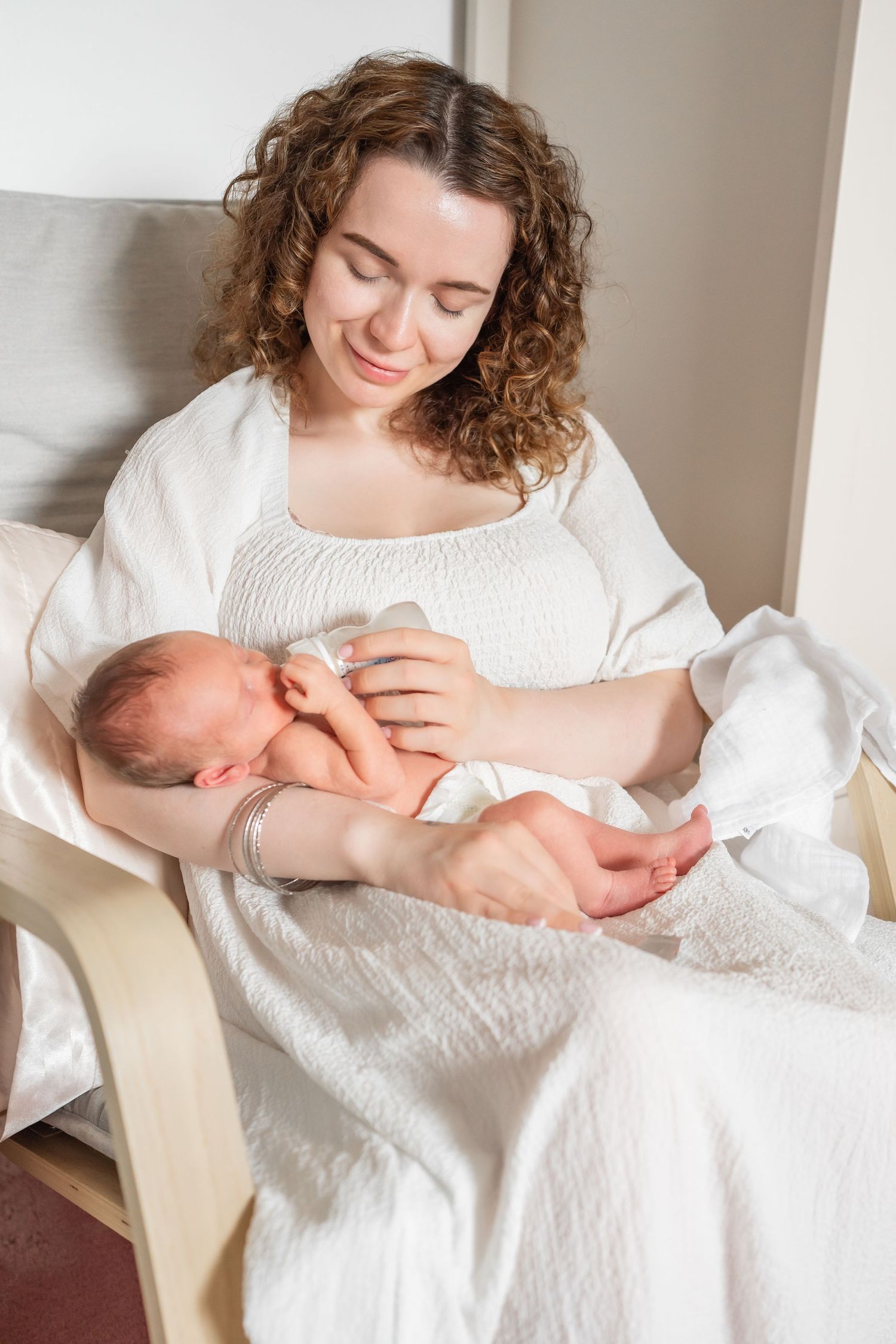 A smiling mother in a white dress sits in a chair cradling her sleeping newborn baby.