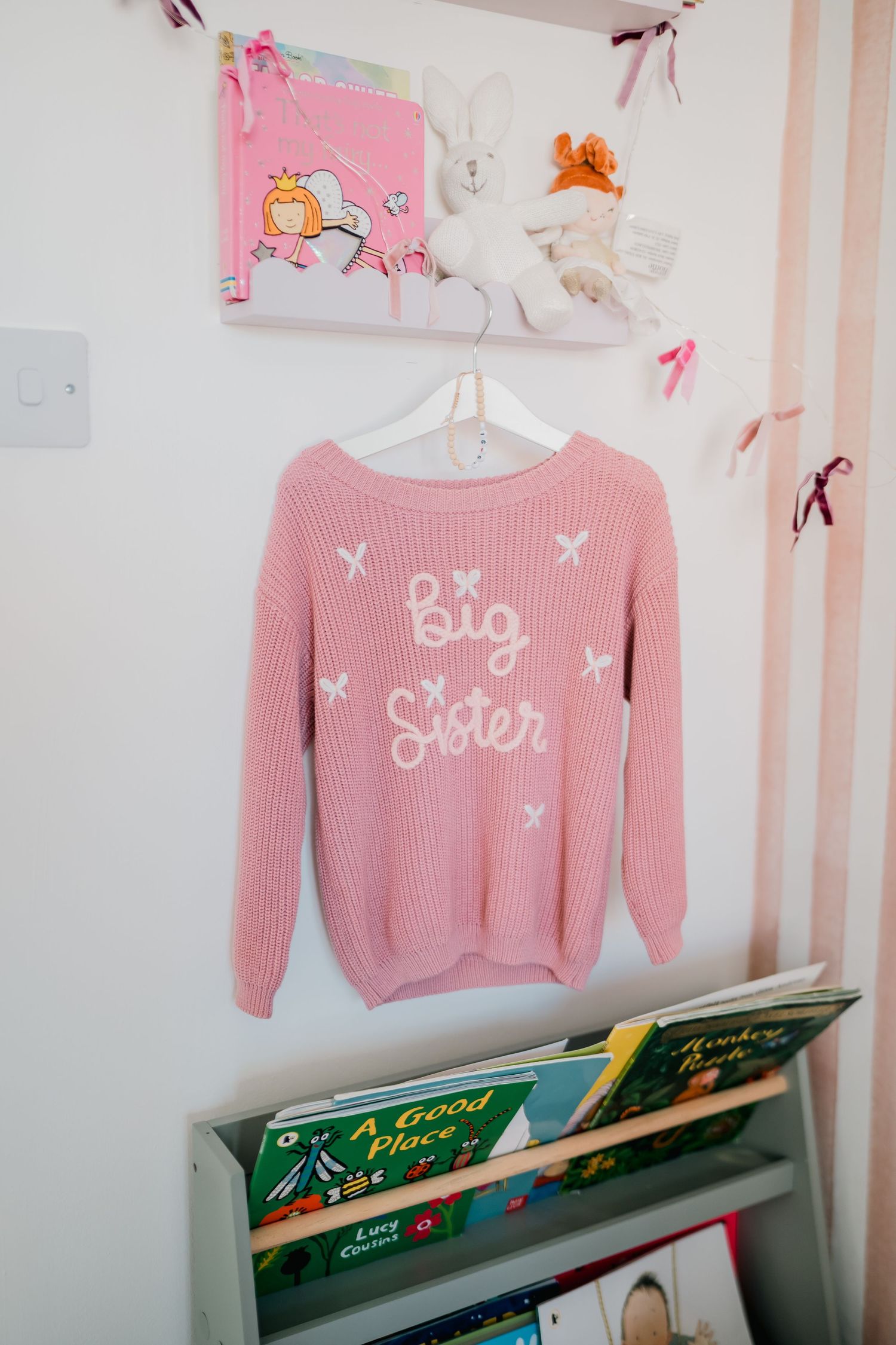 Pink knit Big Sister sweater hanging on wall above children's bookshelf in a girl's nursery room.