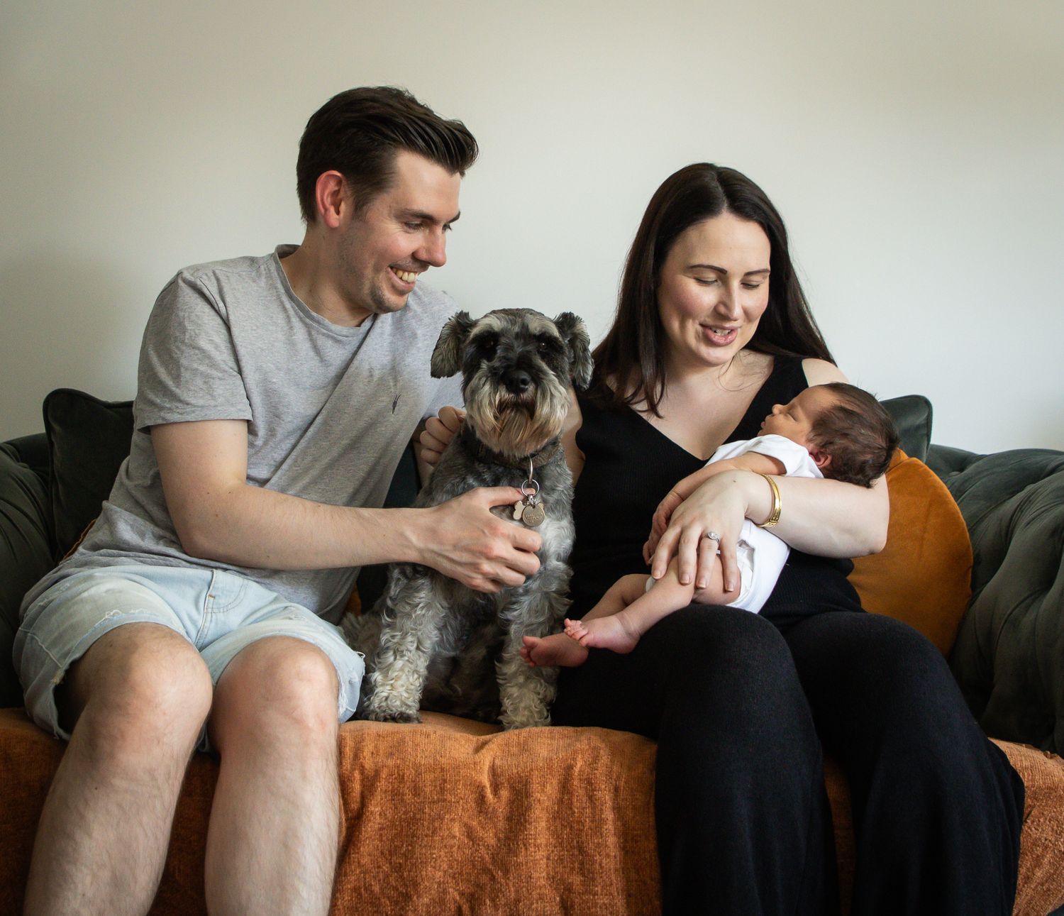 A casual family moment on an orange couch with their dog and infant.