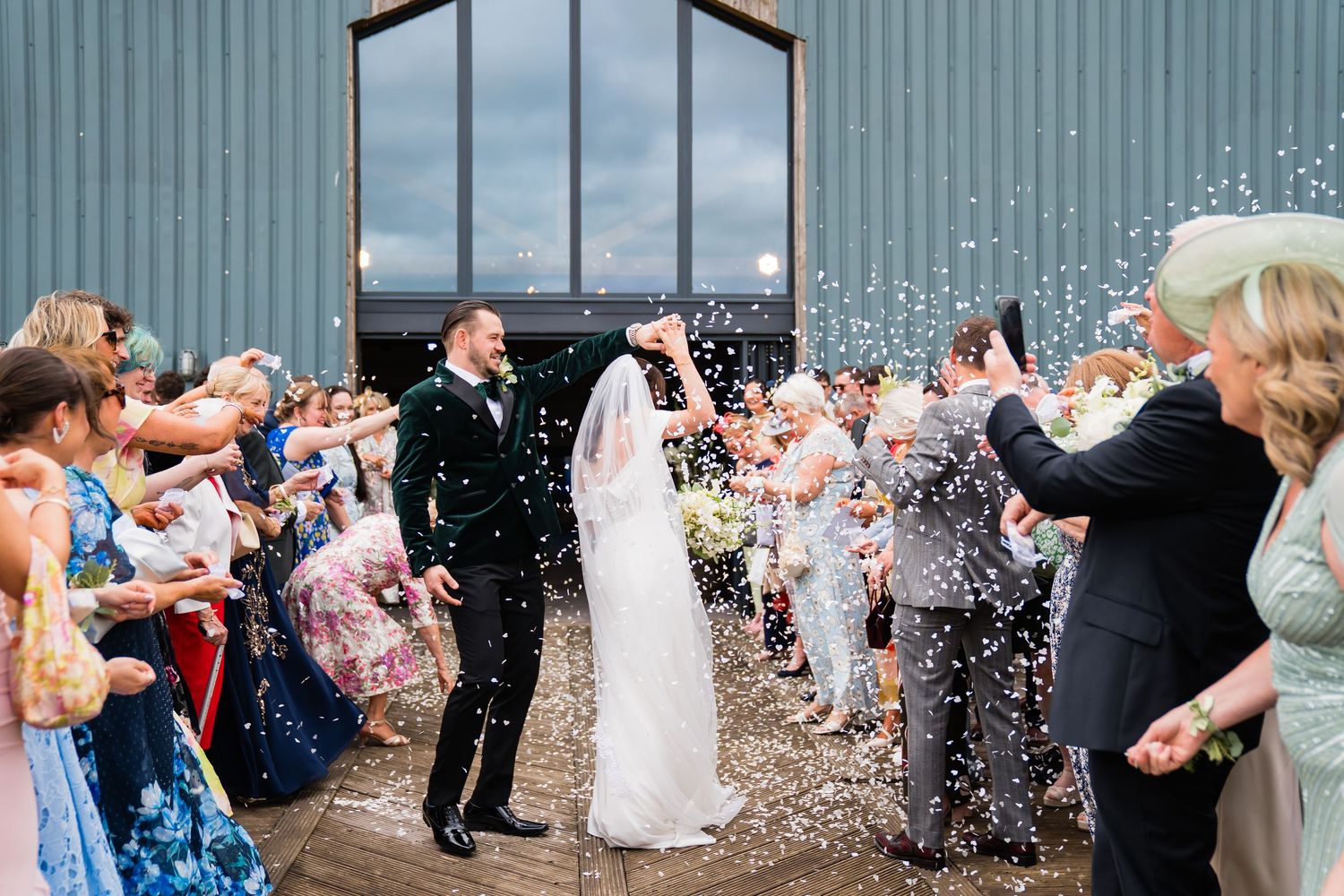 Bride and groom walk through confetti shower outside barn venue, surrounded by cheering wedding guests.