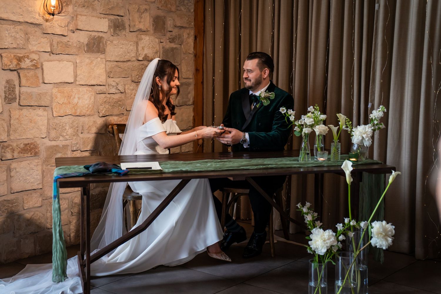 Bride and groom signing wedding register at rustic stone wall venue, surrounded by white floral arrangements.