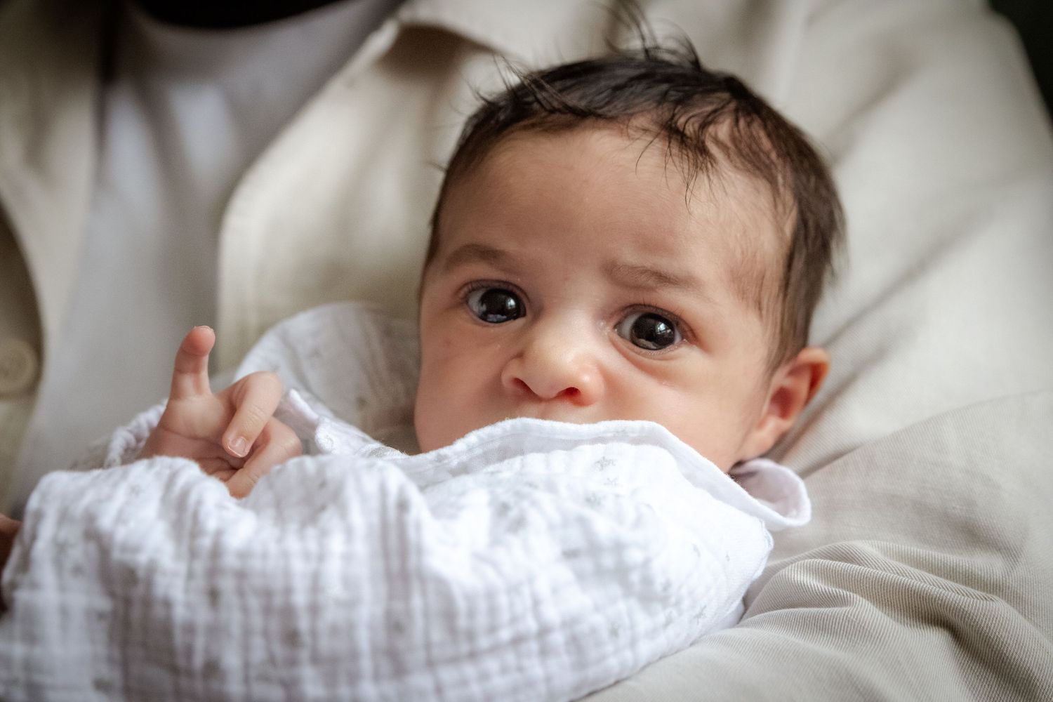 A baby wearing a white muslin swaddle blanket rests on soft bedding.