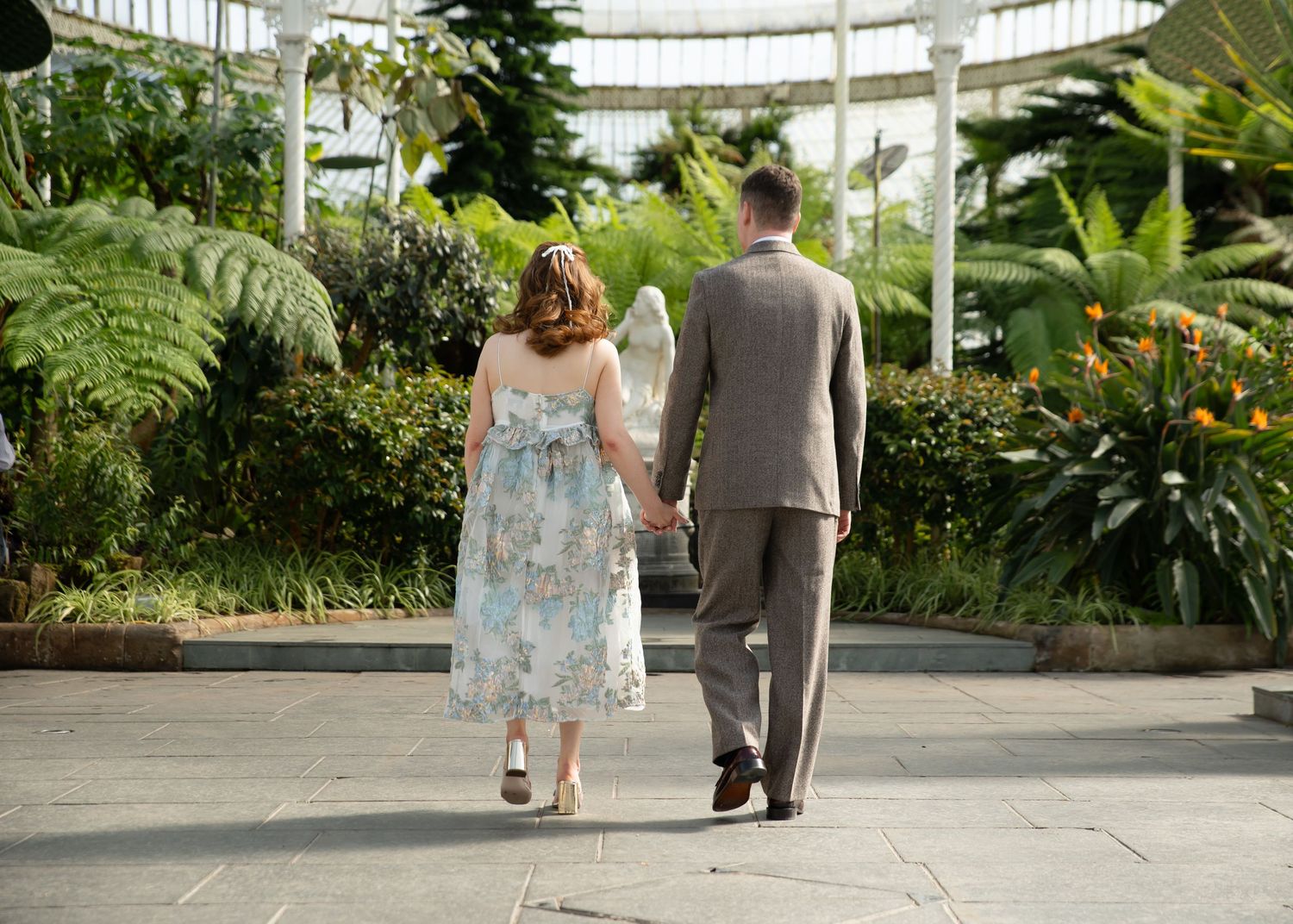 Couple strolling through a lush botanical garden with glass conservatory.