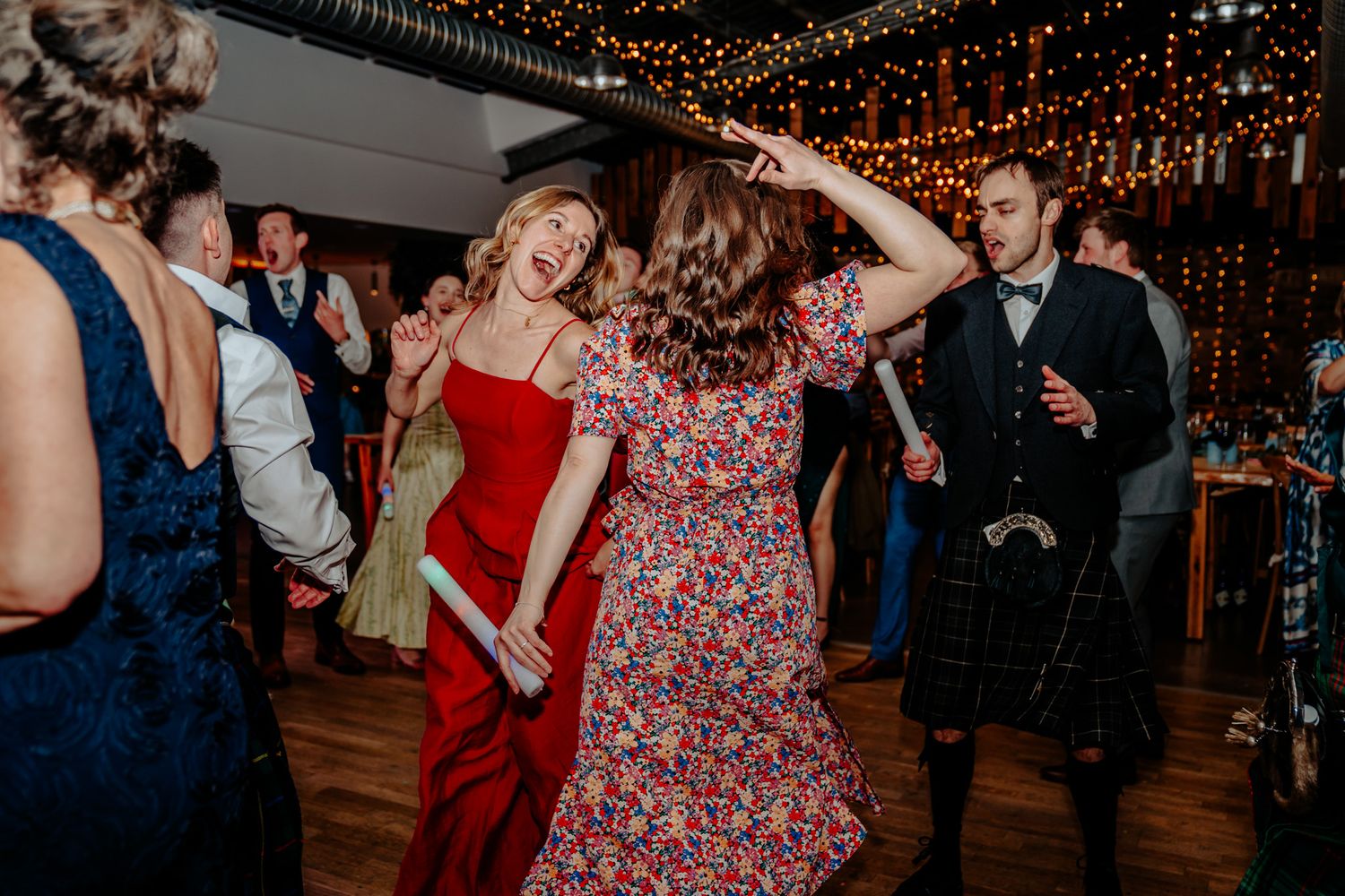 Guests dancing joyfully at a wedding reception, woman in red dress twirling under fairy lights in a rustic venue.