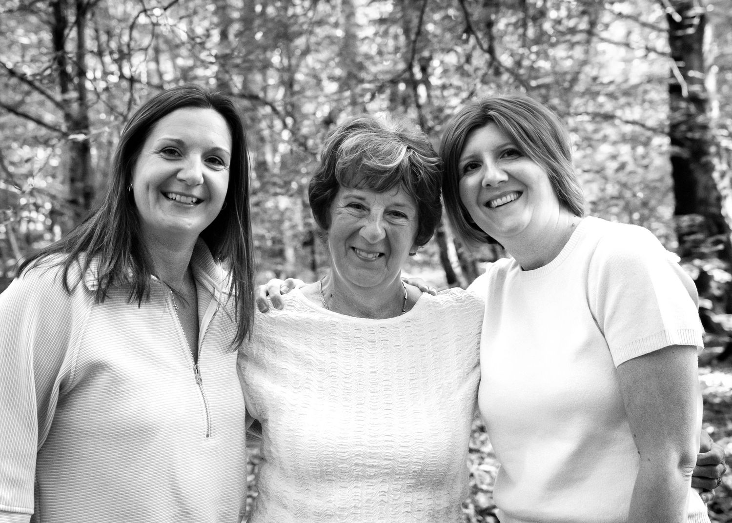 Three smiling women pose together outdoors in a black and white photo among trees.