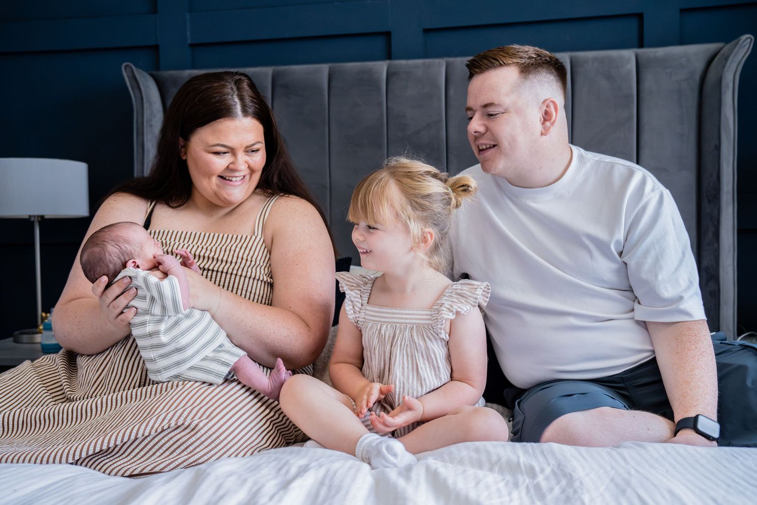 A happy family of four sits on a bed, with mom holding a newborn, dad smiling, and a young daughter between them.