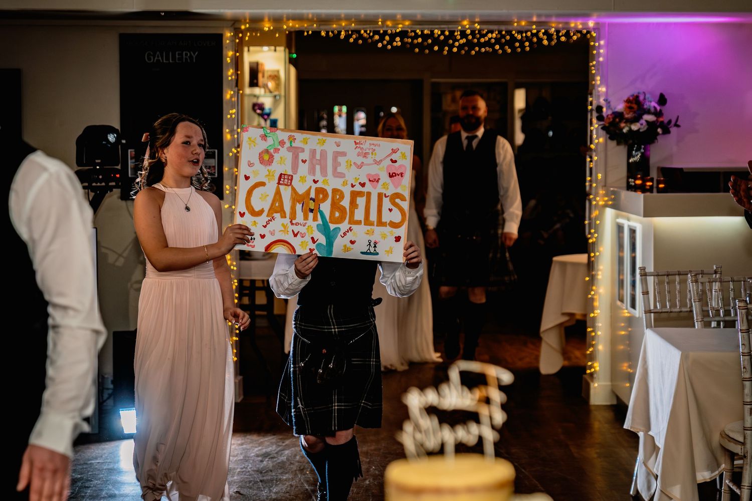 Wedding guests holding a decorative The Campbells sign at a reception venue decorated with fairy lights.