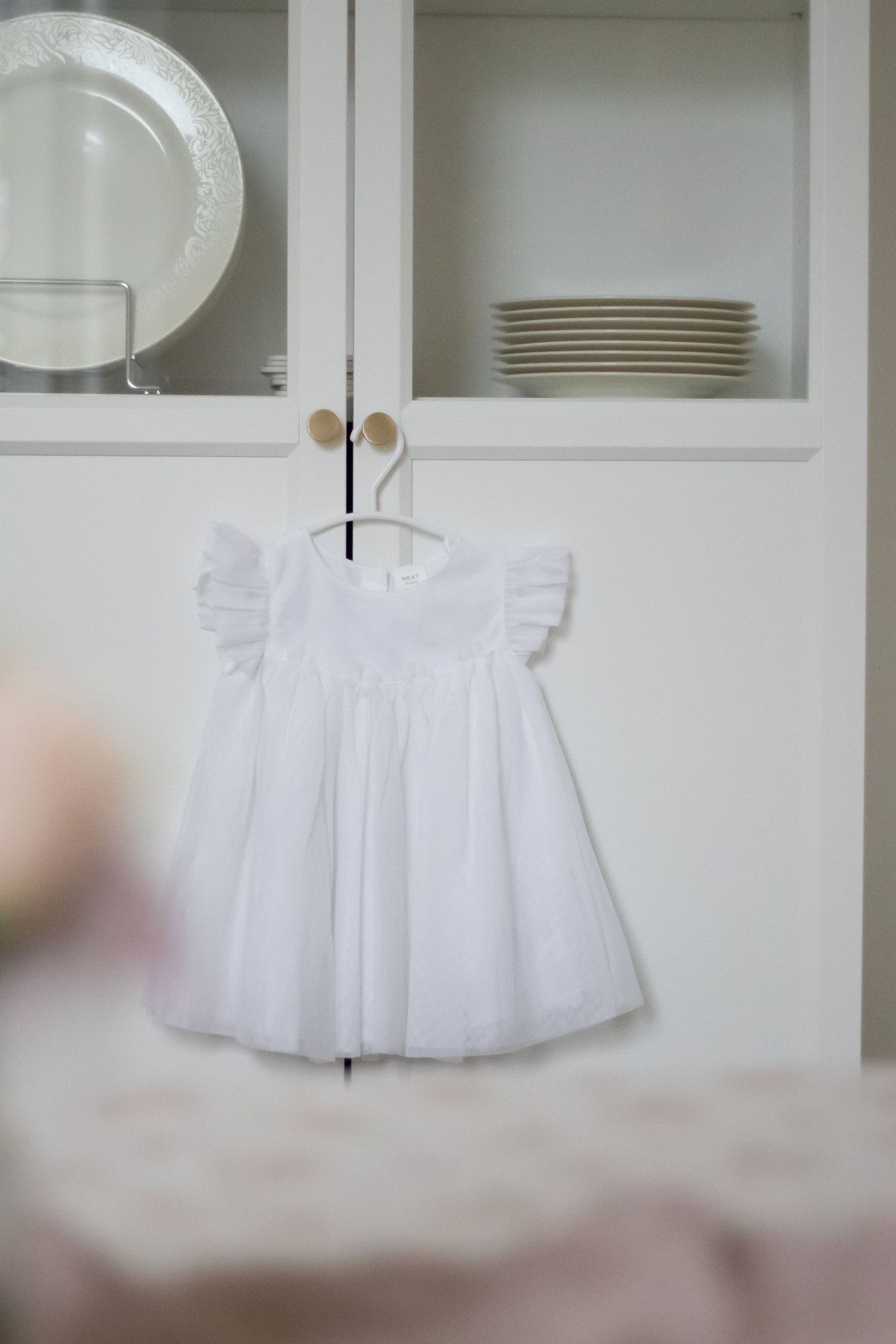 White baby girl dress with flutter sleeves and tulle skirt hanging on a hanger in front of a white cabinet.