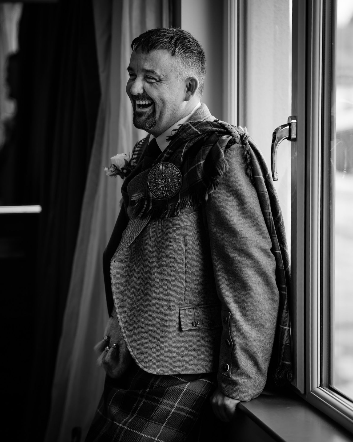 A laughing groom in a Scottish kilt and tweed jacket with a thistle boutonniere stands by a window in a black and white photo.