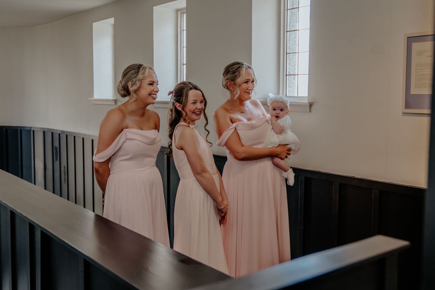 Three bridesmaids in blush pink dresses stand together, one holding a baby, smiling at a wedding venue.